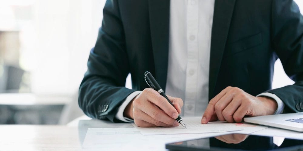 Seated man is suit reviewing paperwork with a pen in his hand