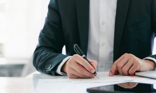 Seated man is suit reviewing paperwork with a pen in his hand